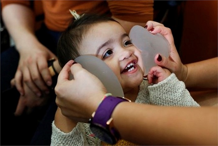 Hình ảnh một bé gái đang đùa nghịch với túi ngực silicon trong khi mẹ của cô bé đang được bác sỹ kiểm tra y tế.