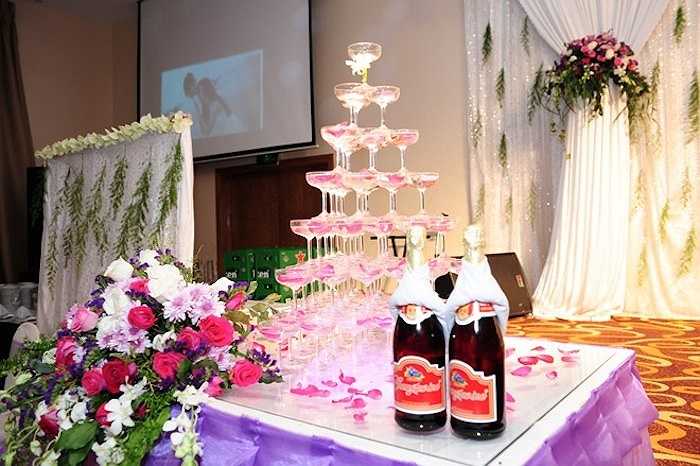 Không gian tiệc bên trong có gam màu trắng, tím, hồng ngọt ngào. Tháp rượu đặt ngay ngắn được trang trí bởi những cánh hoa hồng. 