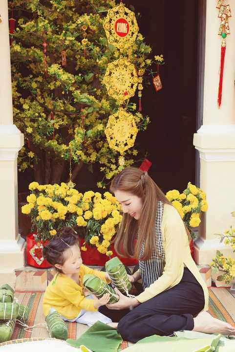 Riêng bé Cadie Mộc Trà, với trang phục tông xuyệt tông cùng mẹ, khoe vẻ đáng yêu, nụ cười tươi tắn như thiên thần. Cô bé còn cùng mẹ thực hiện những động tác gói bánh Tét hay cảnh bé trao cho mẹ quả dưa đầy giữa không gian ngập sắc vàng của hoa mai phương Nam.