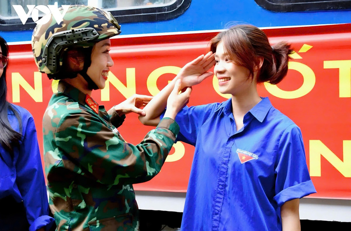 Trong khoảnh khắc đầy ý nghĩa tại ga Đà Nẵng, một nữ chiến sĩ tận tình chỉnh sửa động tác chào cờ cho nữ đoàn viên thanh niên. Nụ cười tươi tắn, ánh mắt tràn đầy sự động viên và tự hào thể hiện tình cảm gắn bó giữa những người trẻ.