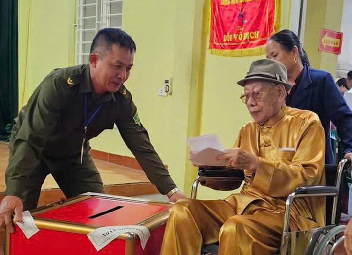 Một cử tri cao tuổi phải ngồi xe lăn ở phường Hạc Thành (Thanh Hoá) được thành viên tổ bầu cử hỗ trợ bỏ phiếu. (Ảnh: Công an Thanh Hoá)