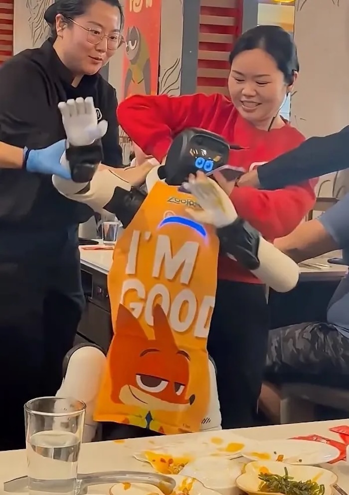 Robot mặc chiếc tạp dề màu cam với dòng chữ “Tôi ổn”, vừa lắc tay vừa lắc hông theo điệu nhạc.