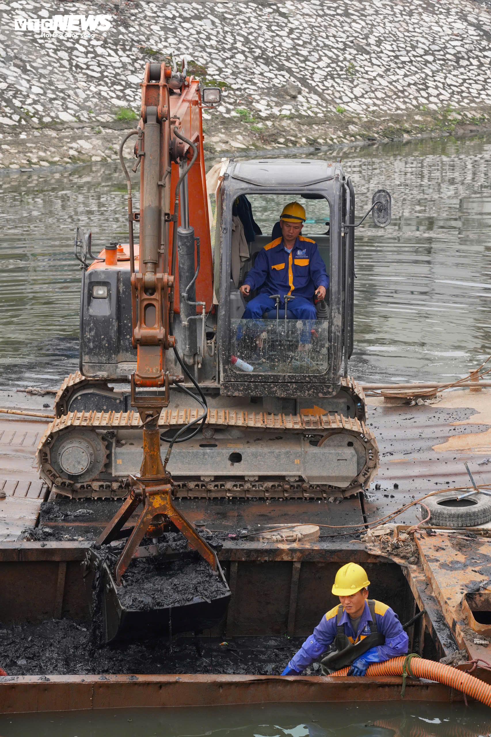 Dưới lòng sông Kim Ngưu, một máy xúc gầu ngoạm được bố trí trên phao nổi để múc bùn. Bên cạnh đó là thuyền sắt phục vụ công nhân tiếp cận khu vực thi công. Một công nhân trực tiếp giữ ống hút dưới nước, phối hợp với tổ vận hành trên bờ để hút bùn thải vào các xe téc chờ sẵn, đưa đi xử lý.